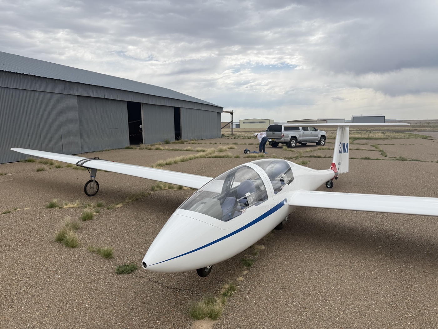 A white two-seat glider parked outside its hangar.